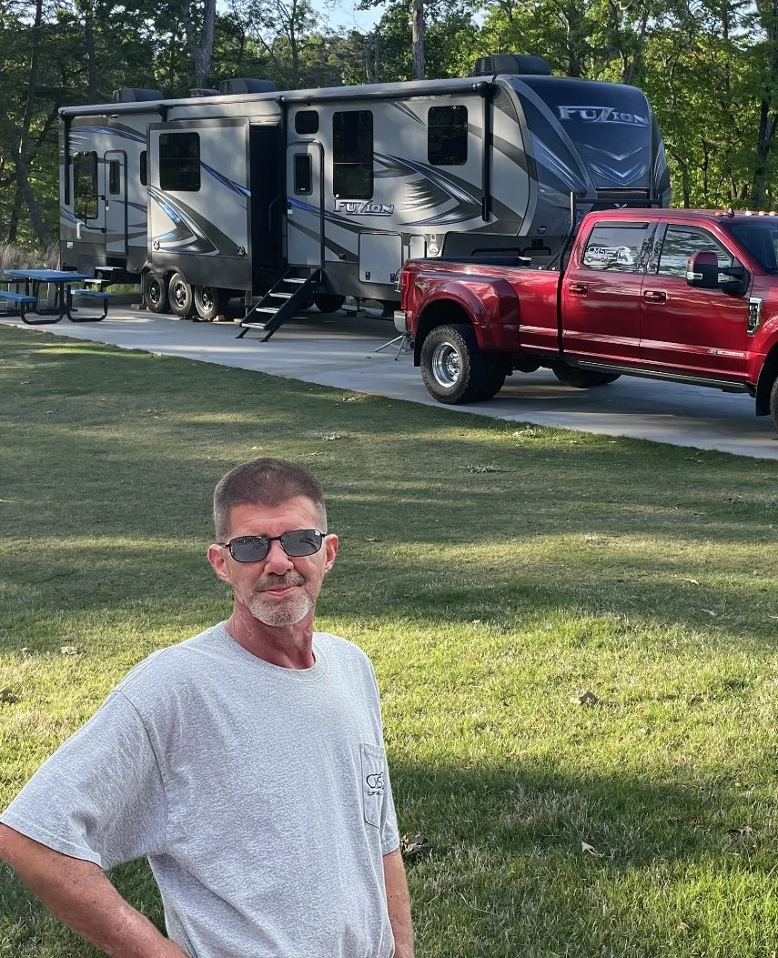 A man wearing sunglasses and a light gray t-shirt standing outdoors on grass, with a red pickup truck and a large gray modern RV parked on a paved area behind him surrounded by trees.
