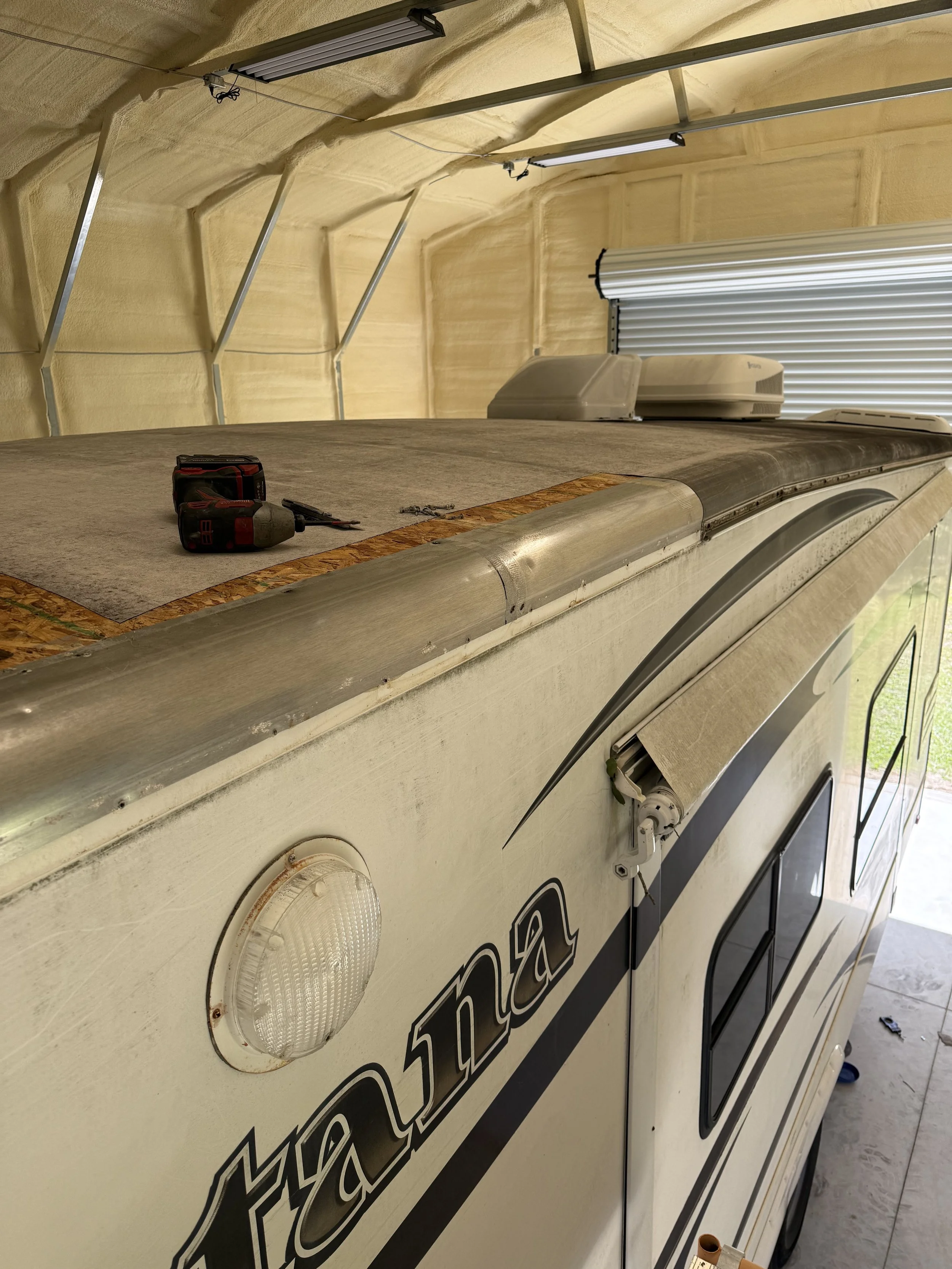 Close-up of the side of a white camper van with black lettering, two round white lights, and some tools placed on top, inside a garage with yellow insulation and fluorescent lights overhead.