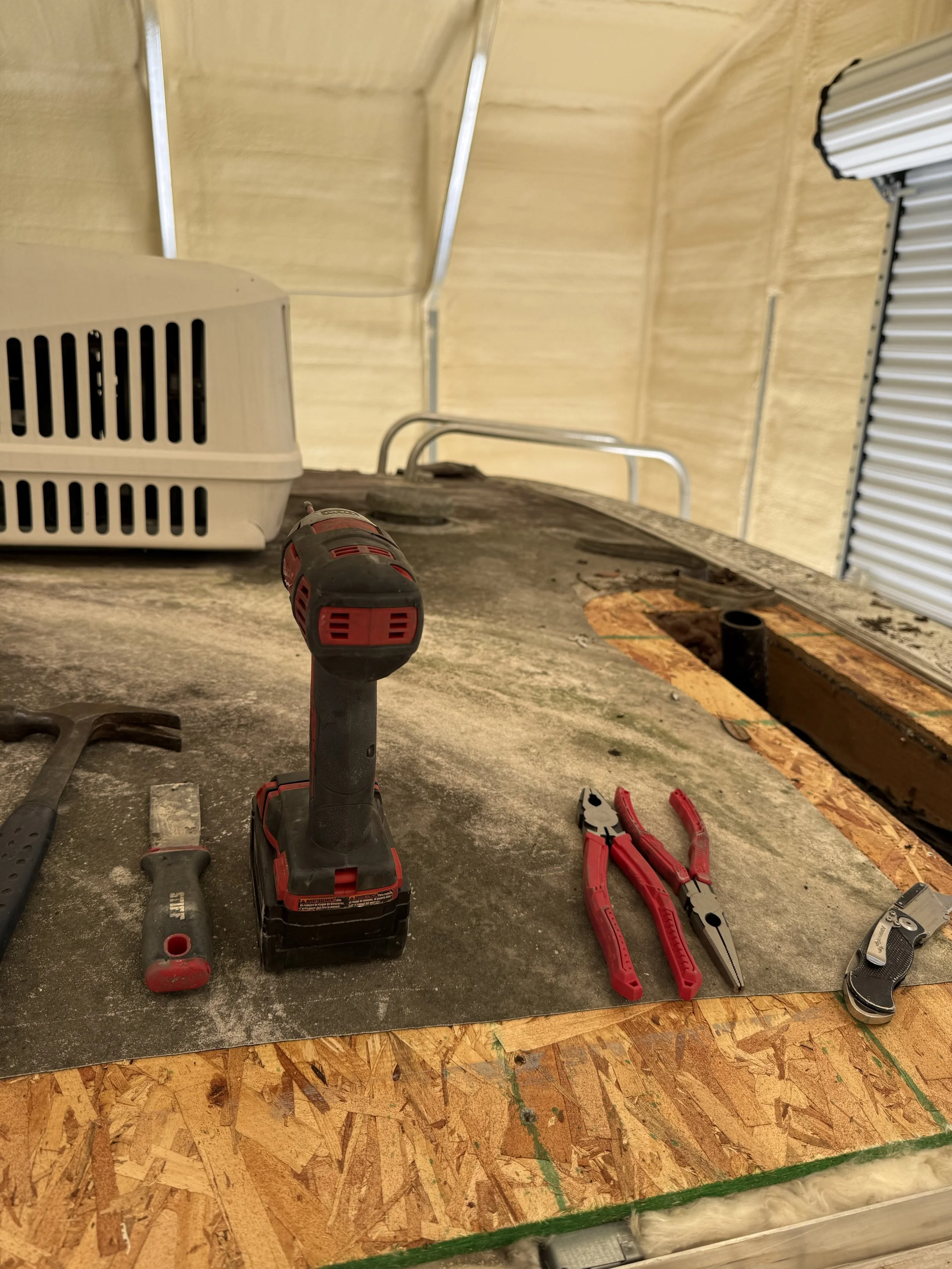 Tools including a cordless drill, pliers, and screwdrivers are placed on a work surface. There is an open space in the wooden platform, and a caulking gun is nearby. The setting appears to be a workshop or construction area.