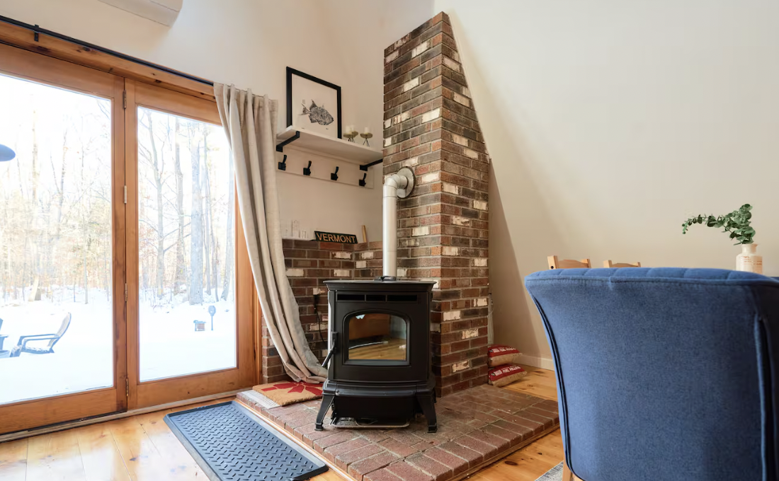 Indoor room with wood flooring, a black wood stove on a brick platform, a brick chimney, a sliding glass door with curtains leading outside to a snowy landscape, and a blue armchair with a plant on a side table.