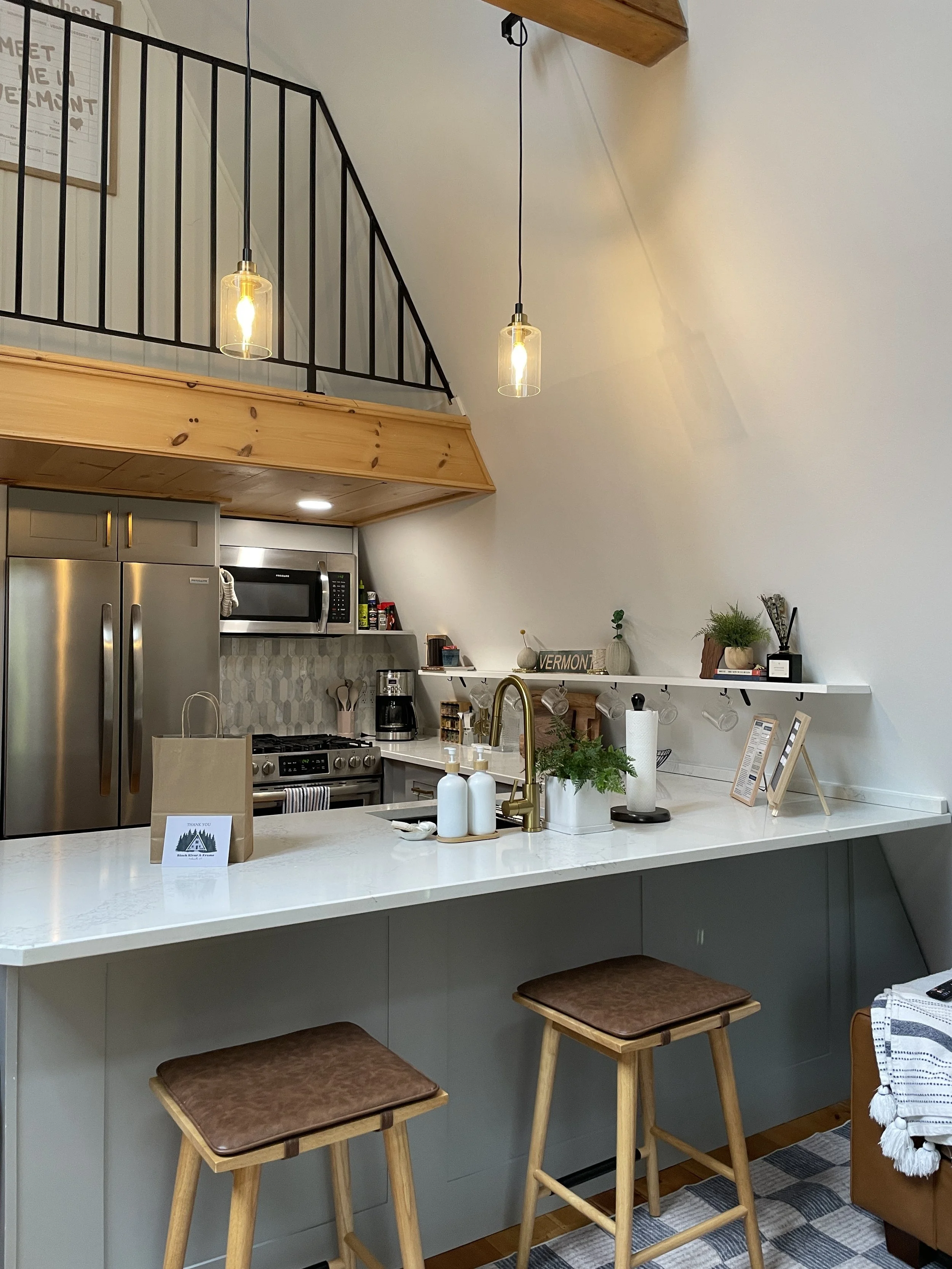 Kitchen with a white countertop, three wooden stools with brown leather seats, stainless steel refrigerator, microwave, coffee maker, and shelves with decorative items and plants. Two hanging pendant lights illuminate the space.