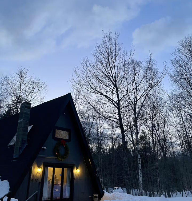 A black A-frame house with a wreath hanging near the porch, illuminated by two yellow porch lights, surrounded by snow and leafless trees under a cloudy sky.