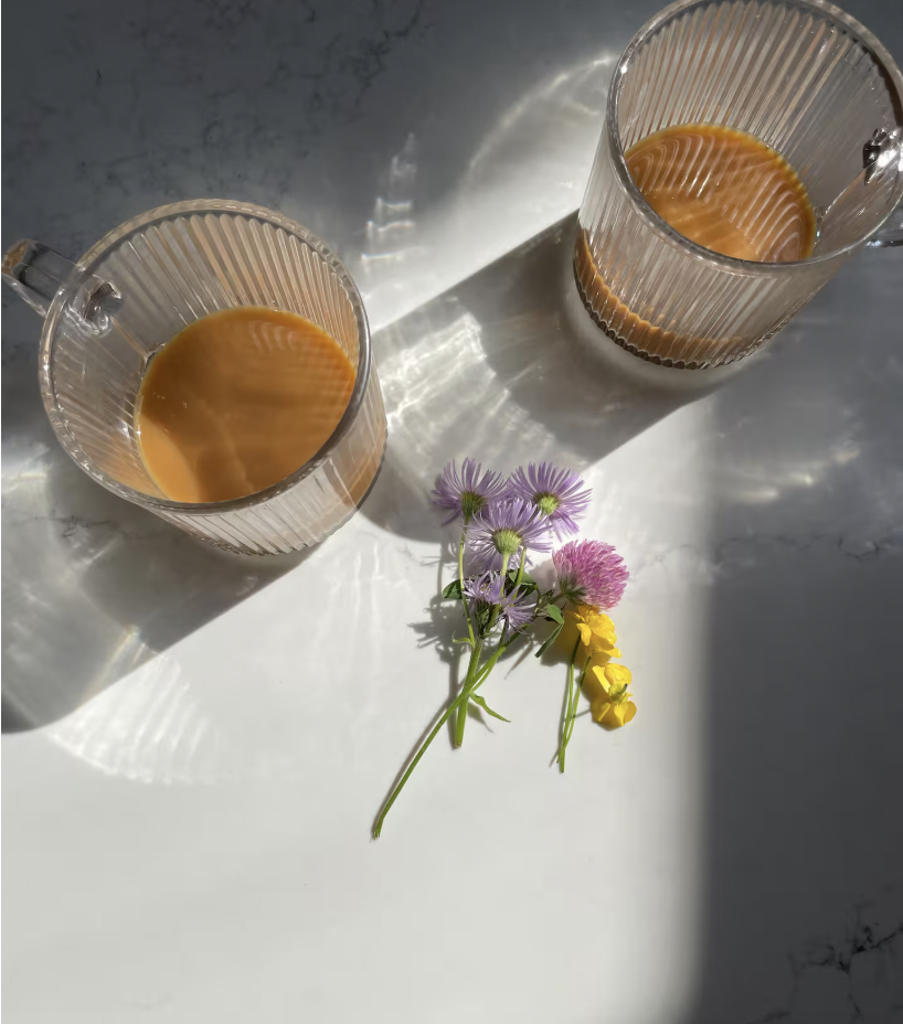 Two glass mugs with textured vertical lines on a marble surface, filled with coffee, arranged with a small bouquet of purple, pink, and yellow flowers in front of them. Sunlight creates streaks and shadows on the surface.