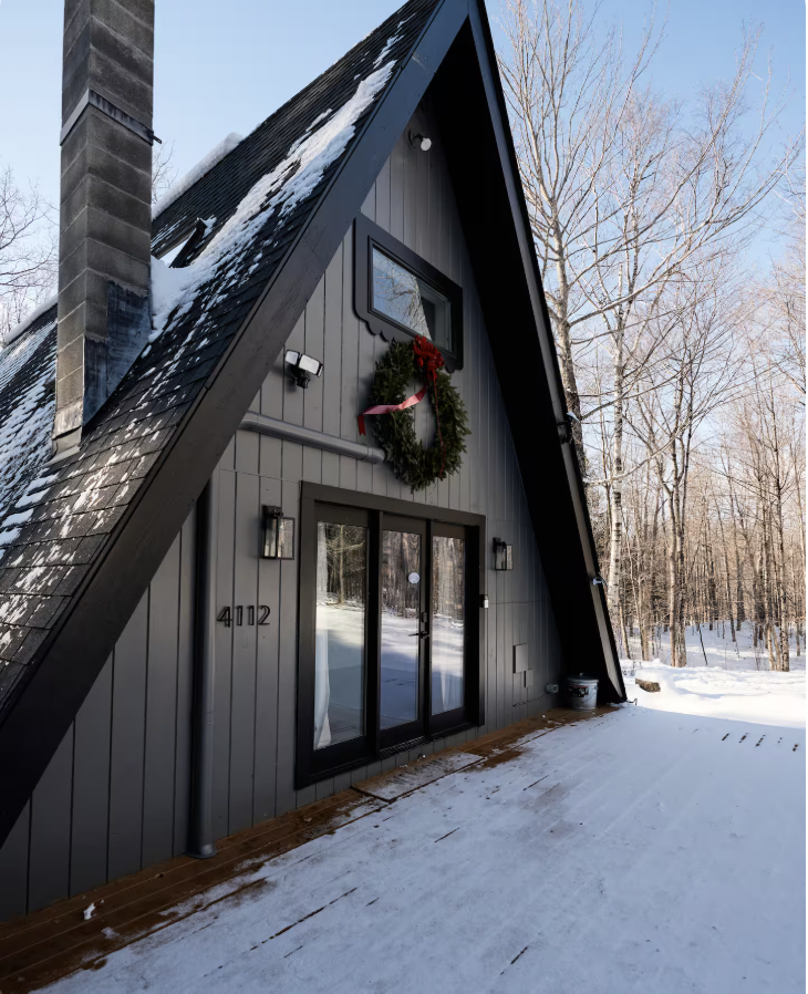 A modern A-frame house with dark gray exterior, decorated with a Christmas wreath, surrounded by snow and leafless trees.