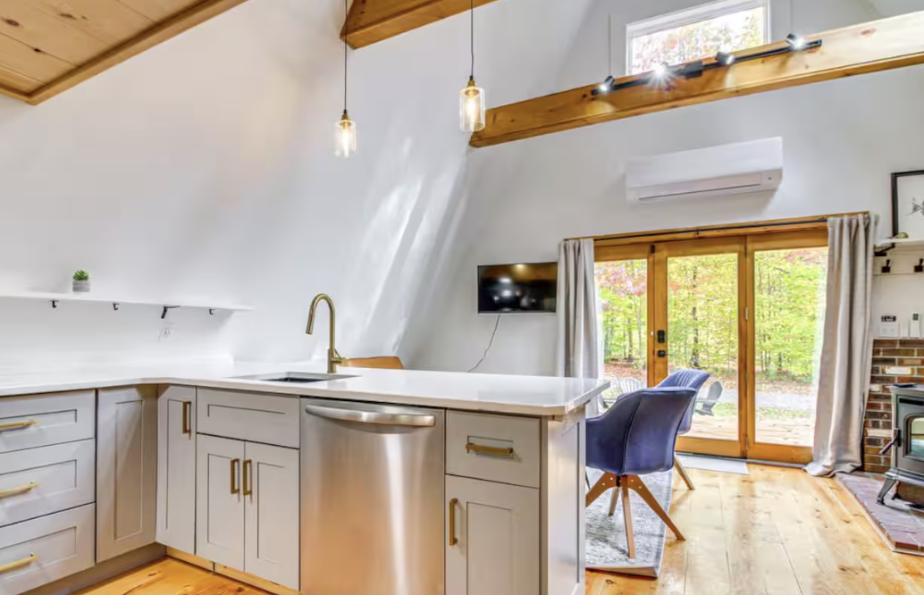Open-concept kitchen with stainless steel dishwasher. Two pendant lights hang from the ceiling. There are sliding glass doors with curtains leading outside to a wooded area. A small TV is mounted on the wall, and a blue chair is in front of the doors