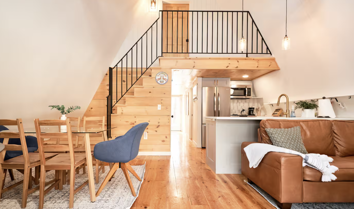 Open-concept living and dining area with wood staircase and kitchen. The dining table has wooden chairs and a blue accent chair, and the living room has a brown leather sofa with pillows and a throw blanket.