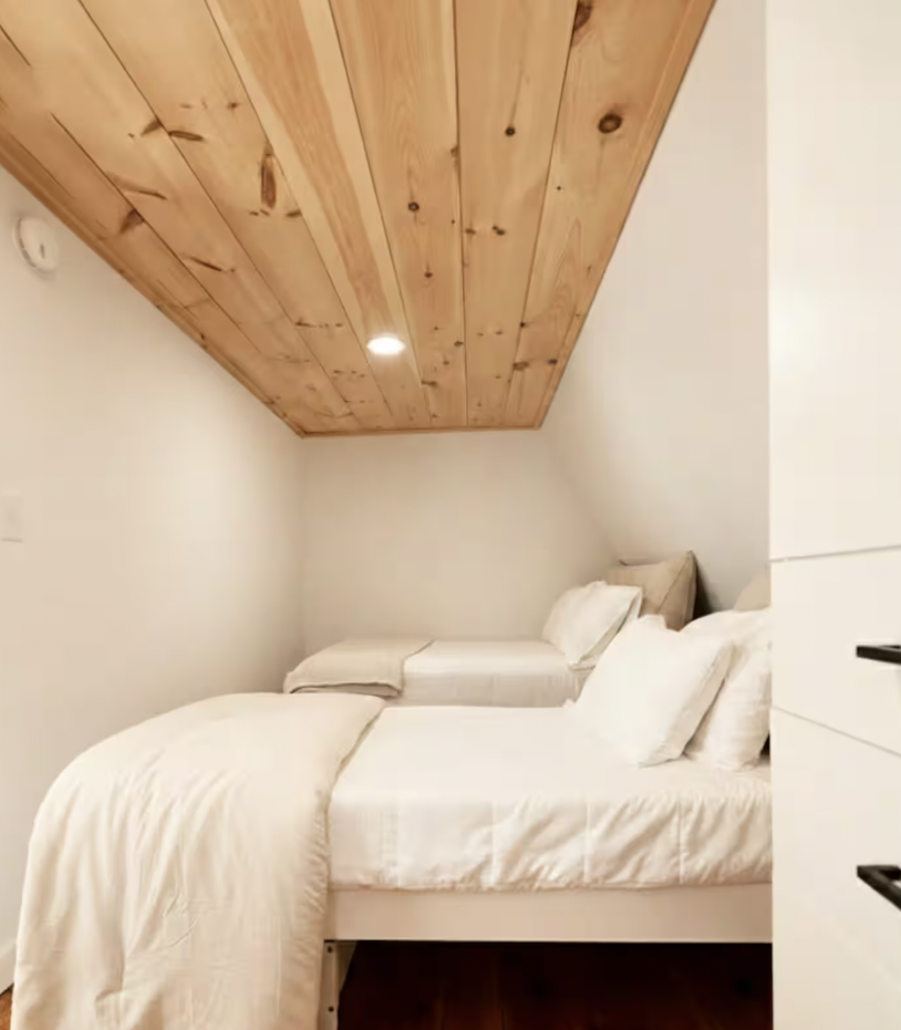 A cozy bedroom with a sloped wood-paneled ceiling, two beds with white bedding and pillows, and neutral-colored walls.
