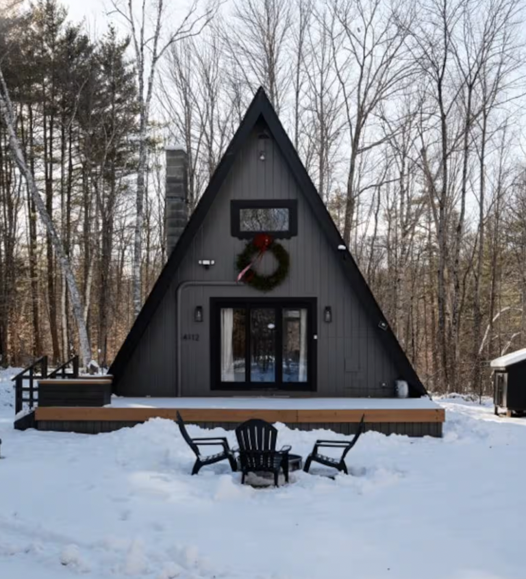 A black A-frame cabin with a sliding glass door, decorated with a Christmas wreath, outdoor seating, and surrounded by snow and trees.