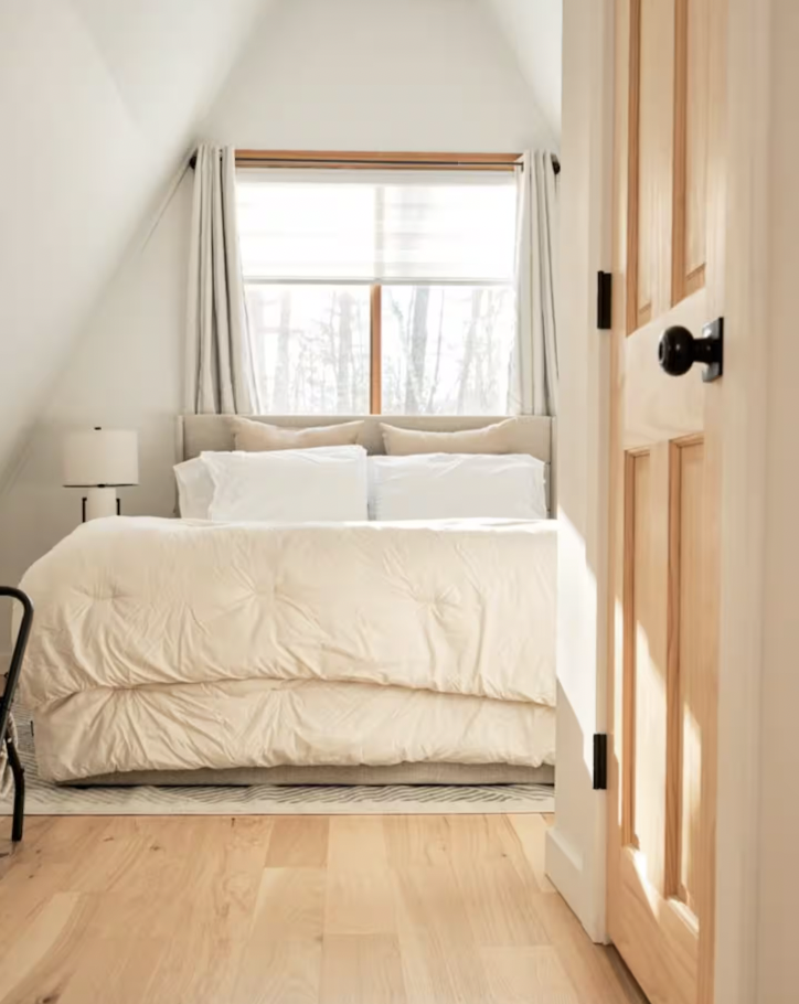Cozy loft bedroom with a queen-sized bed, white bedding, and luxurious pillows, featuring a sloped ceiling with a view of the backyard and woods.