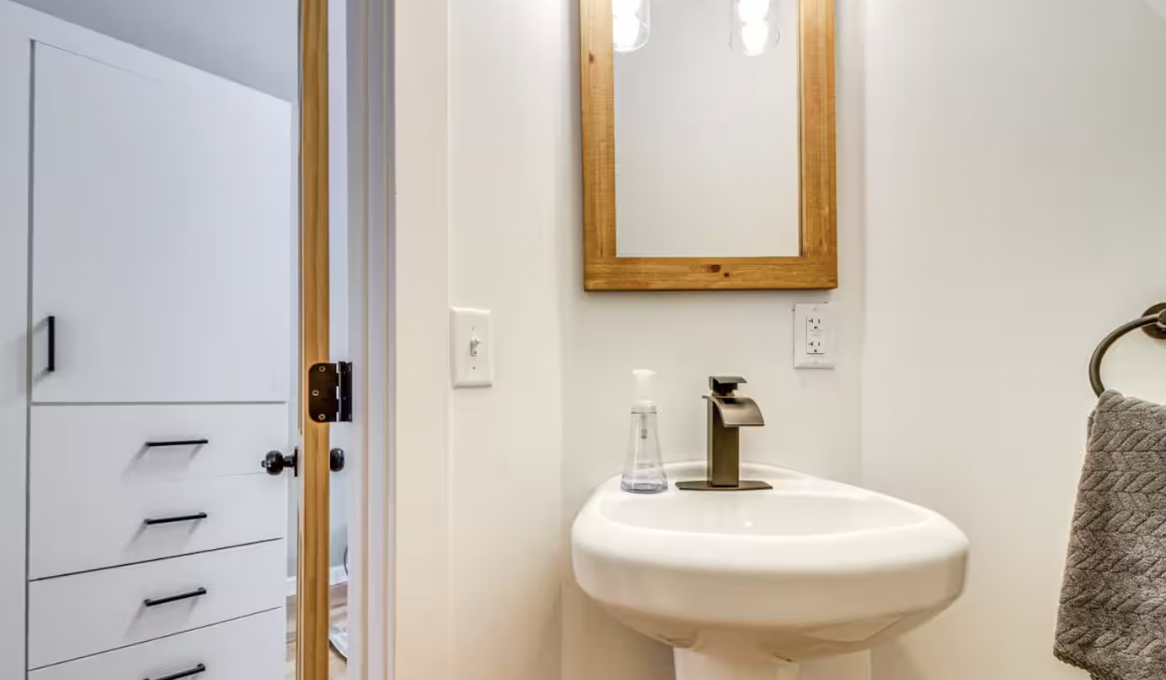 Small bathroom with a white wall-mounted sink, a wooden framed mirror, a towel ring with gray towel, and a cabinet with drawers just outside the doorway.