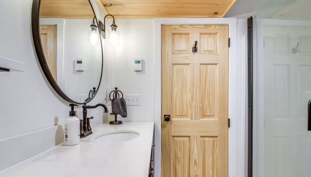 Bathroom with a white countertop, round mirror, black faucet, wall-mounted light fixtures, wooden door, towel holder, and shower area with a glass door.