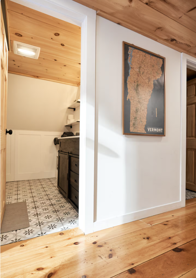 A small bathroom with patterned tile flooring, a wooden vanity, white walls, and open shelves. The ceiling is wood-paneled, and there is a framed map of Vermont on the adjacent wall.