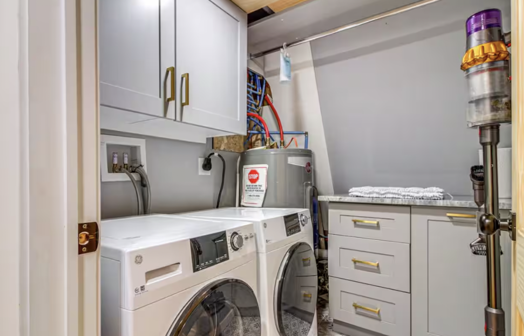 A laundry room with white washer and dryer, white cabinets with golden handles, a countertop, and a cordless vacuum attached to the wall.