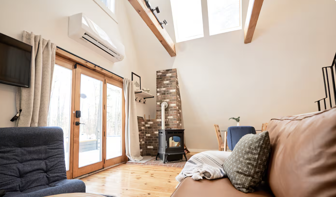Cozy living room with wooden floor, glass sliding doors, a wood stove, brick chimney, a ventless heater, a couch with a pillow and blanket, a chair, a wall-mounted TV, and a high ceiling with skylights.