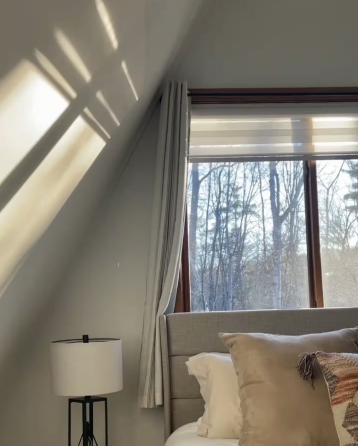 Sunlight creates shadow patterns on a plain wall in a bedroom. A lamp with a white shade is on a black stand next to a bed with beige pillows and a decorative pillow. A window with white and striped curtains shows a view of trees outside.