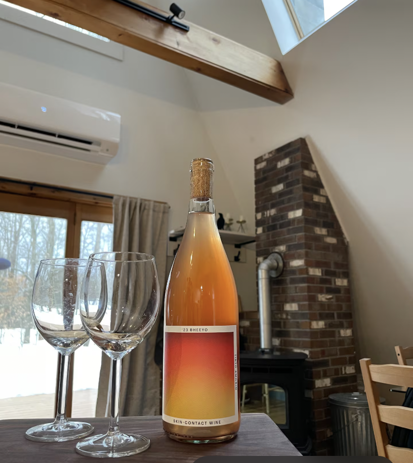 A bottle of skin-contact wine with a gradient label from red to yellow, two empty wine glasses, and a background of a cozy room with a fireplace, wooden beams, and large windows showing a snowy landscape.