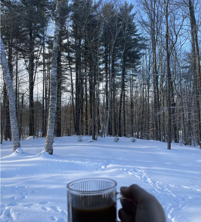 Person holding coffee in front of a snowy forest scene with bare trees and footprints in the snow.