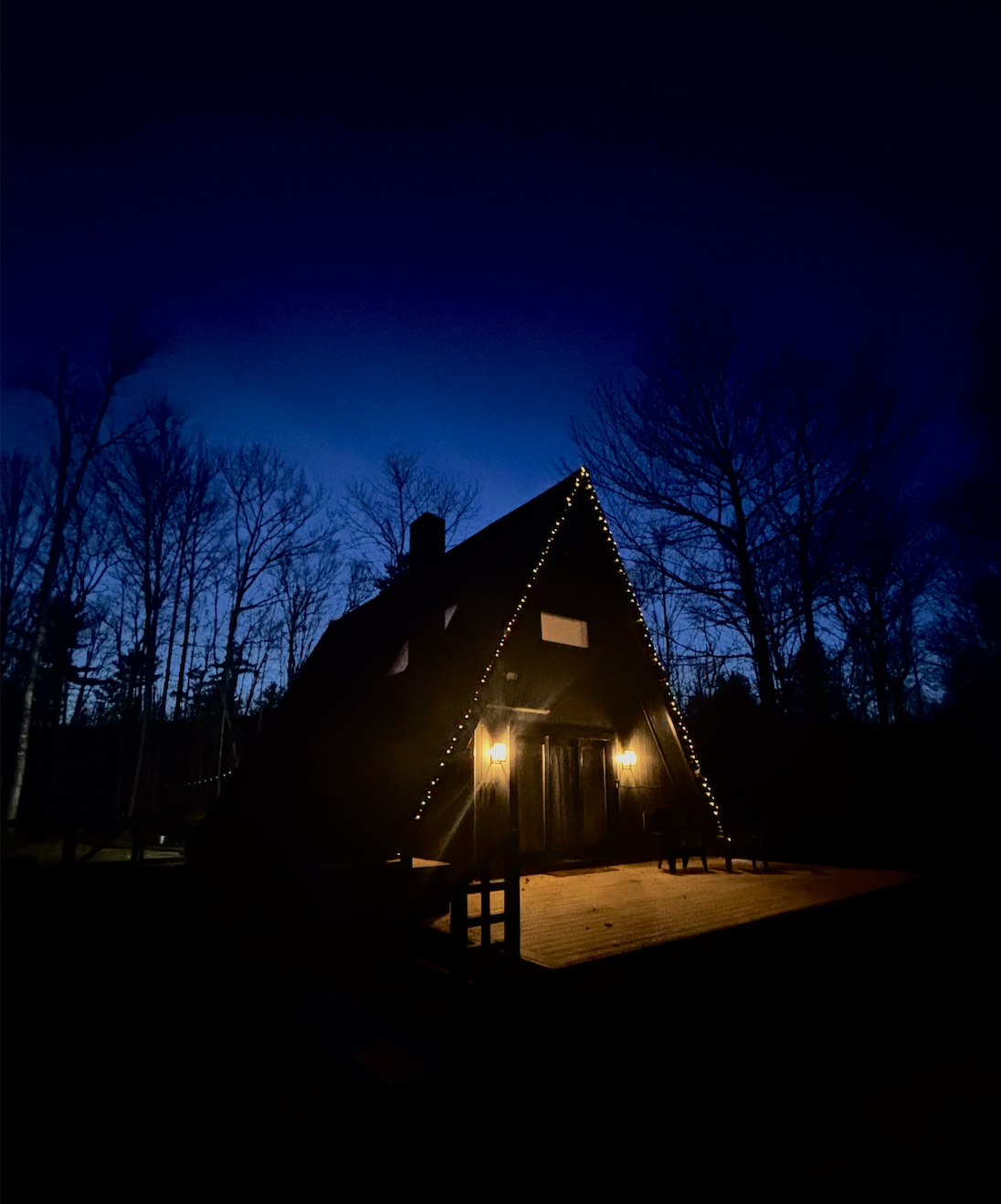 A dark A-frame house decorated with string lights, illuminated from two wall-mounted lamps, surrounded by leafless trees at dusk.