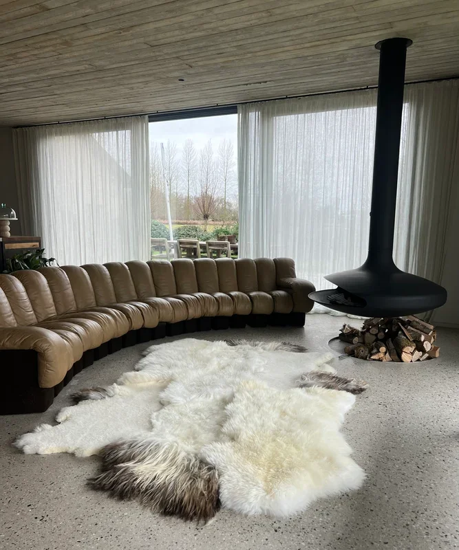 Amy by Wol Society, a creamy white sheep skin rug with grey-brown edges on a speckled floor beside a leather sofa.