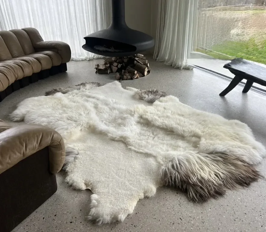 Amy by Wol Society, a creamy white sheep skin rug with grey-brown edges on a speckled floor beside a leather sofa.
