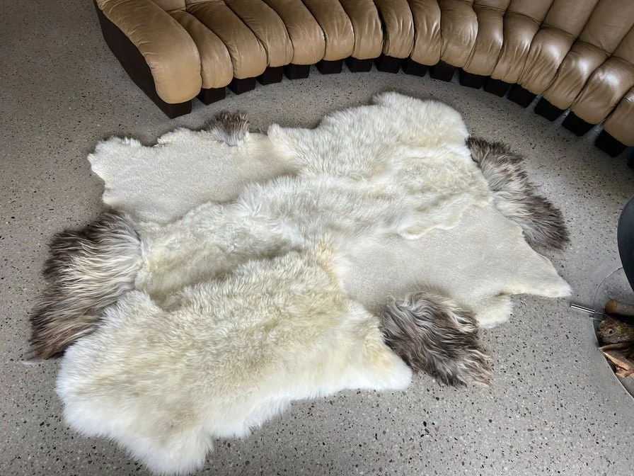 Amy by Wol Society, a creamy white sheep skin rug with grey-brown edges on a speckled floor beside a leather sofa.