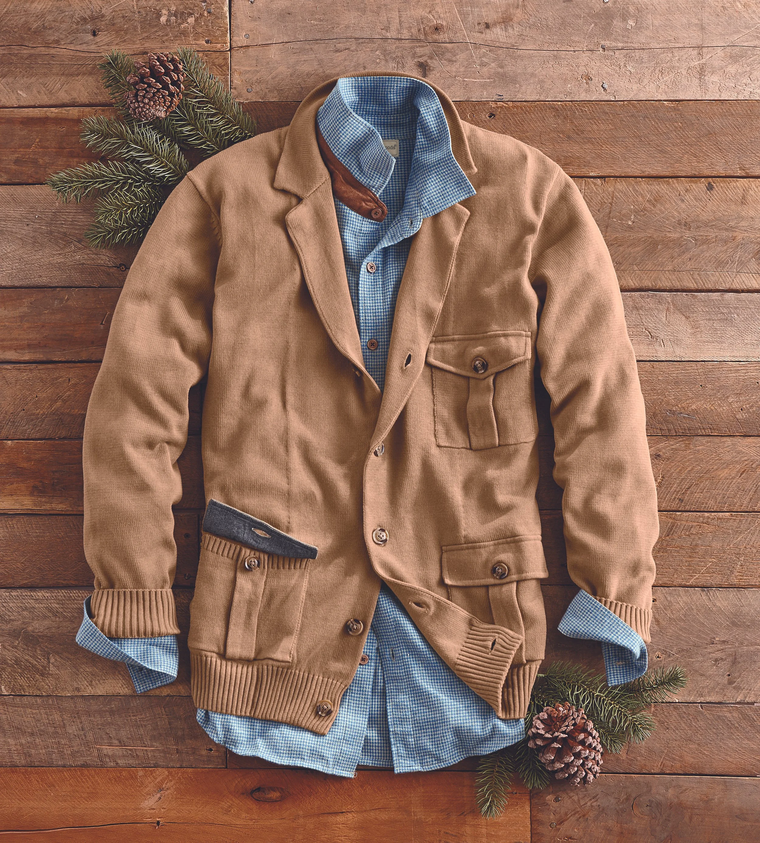 A tan corduroy jacket over a blue and white checked shirt with a brown collar, laid on a wooden surface, decorated with pine cones and evergreen branches.