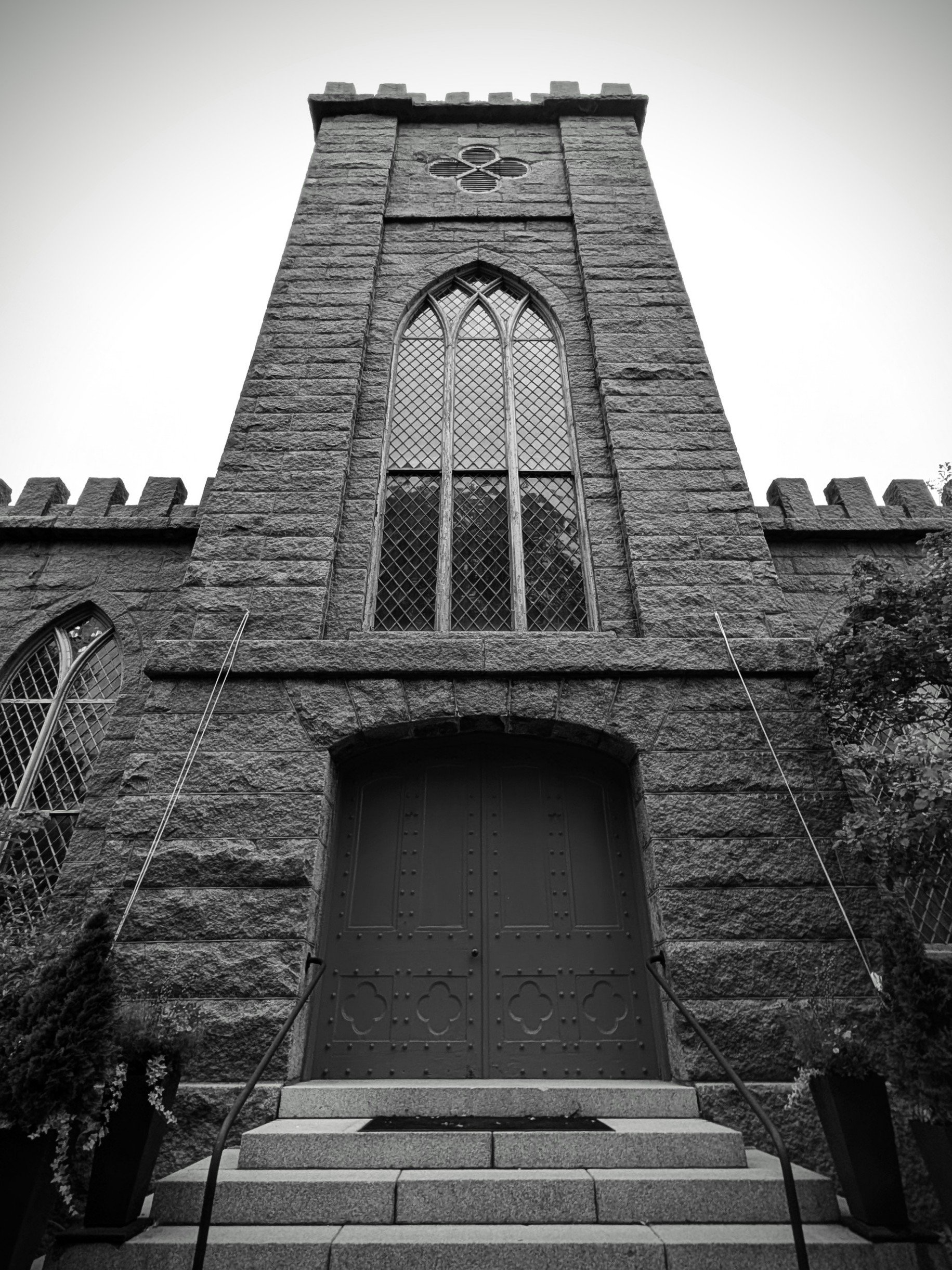 Black and white photo of a stone church with a large gothic arched window and a heavy door, stone steps leading up to the entrance, and two potted plants on either side of the stairs.