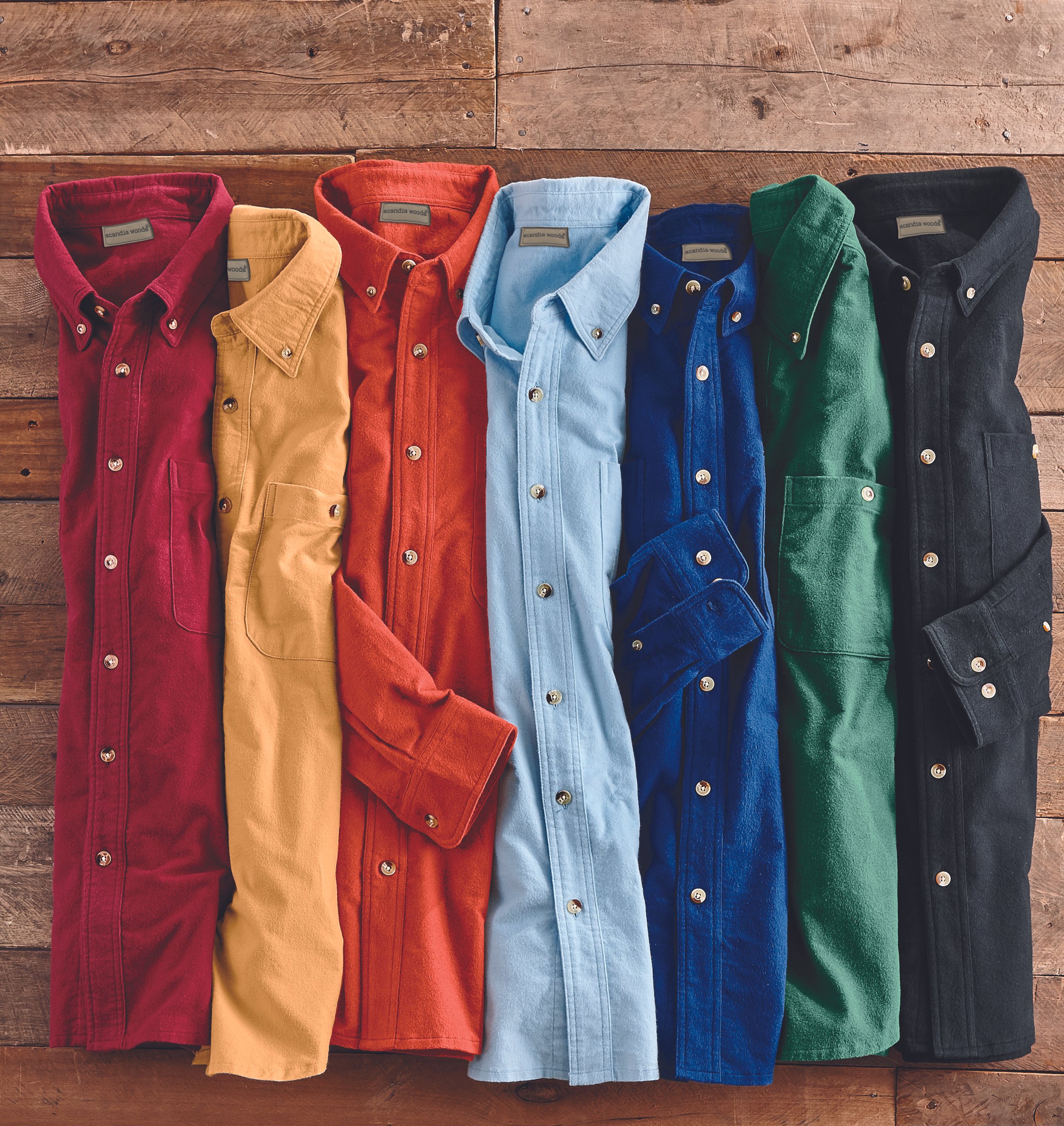 A row of five folded button-up corduroy shirts in red, tan, orange, blue, and black, lying on a wooden surface.