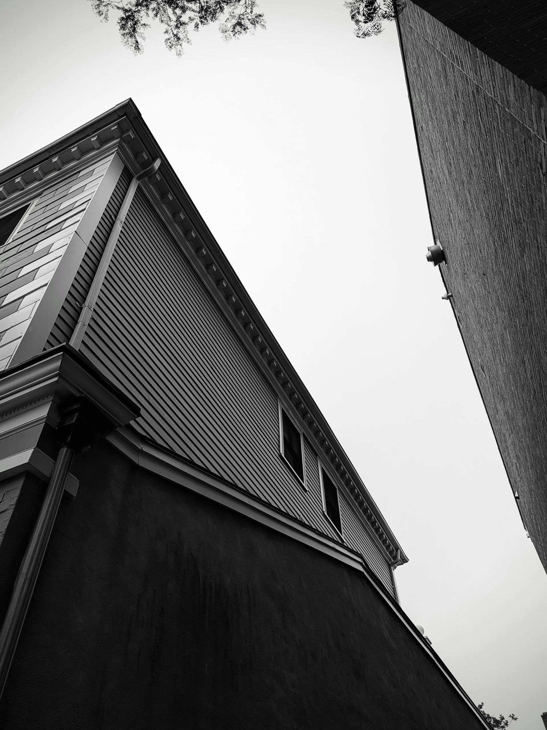 Black and white photo taken from a low angle showing two buildings looking up towards the sky. The building on the left has a storefront with shutters and decorative molding at the top. The building on the right has a textured brick exterior. The sky
