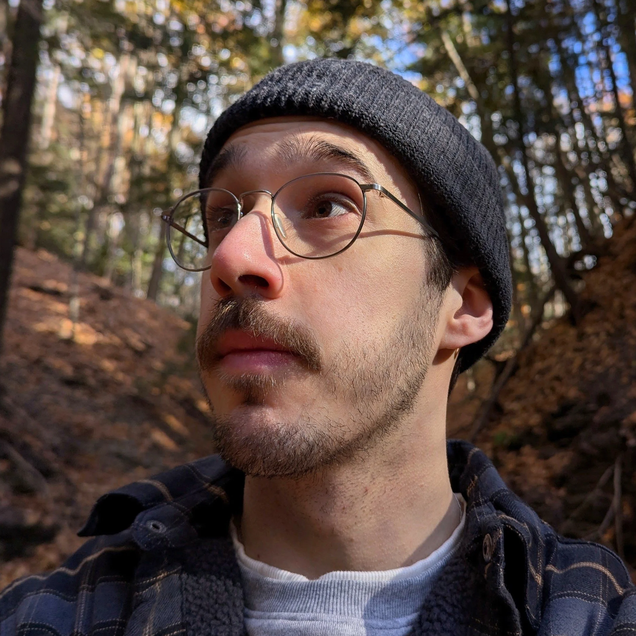 Close-up of a person's face wearing glasses and a black beanie, outdoors in a forest with autumn leaves and sunlight filtering through trees.