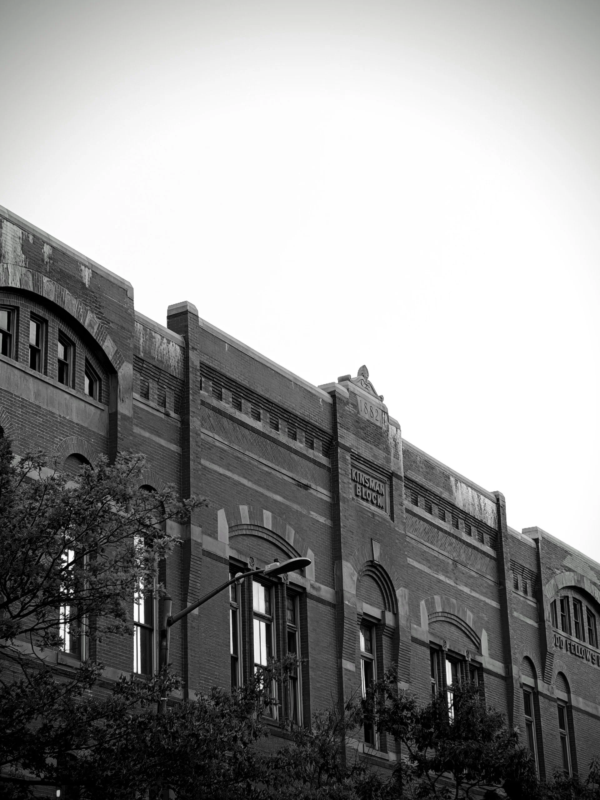 Black and white photo of an old brick building with arched windows and a sign that reads 'Kinsman Block,' with trees in the foreground and the sky in the background.