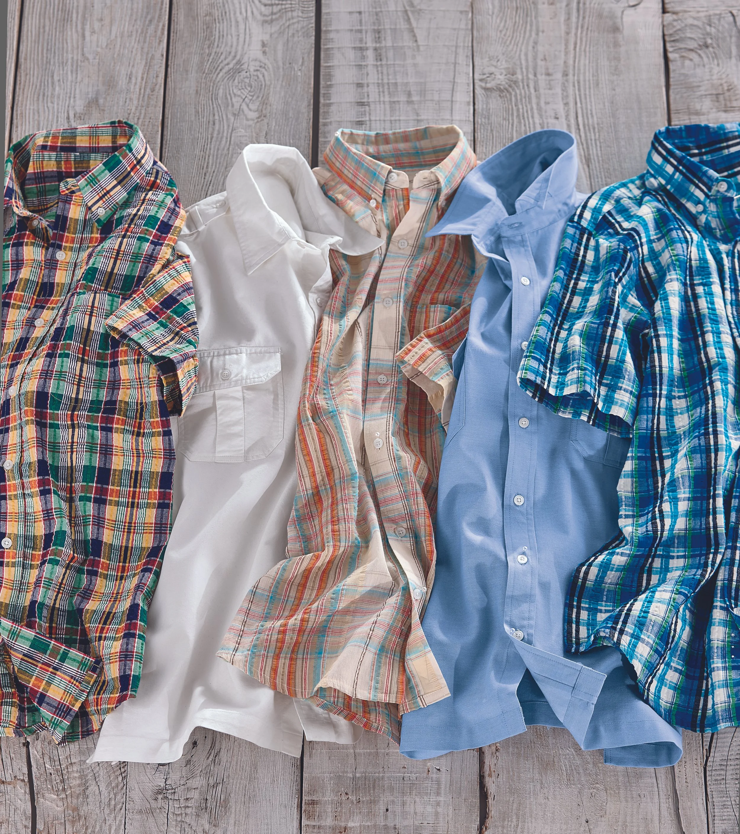 Five plaid and solid color button-up shirts laid out on a wooden surface.