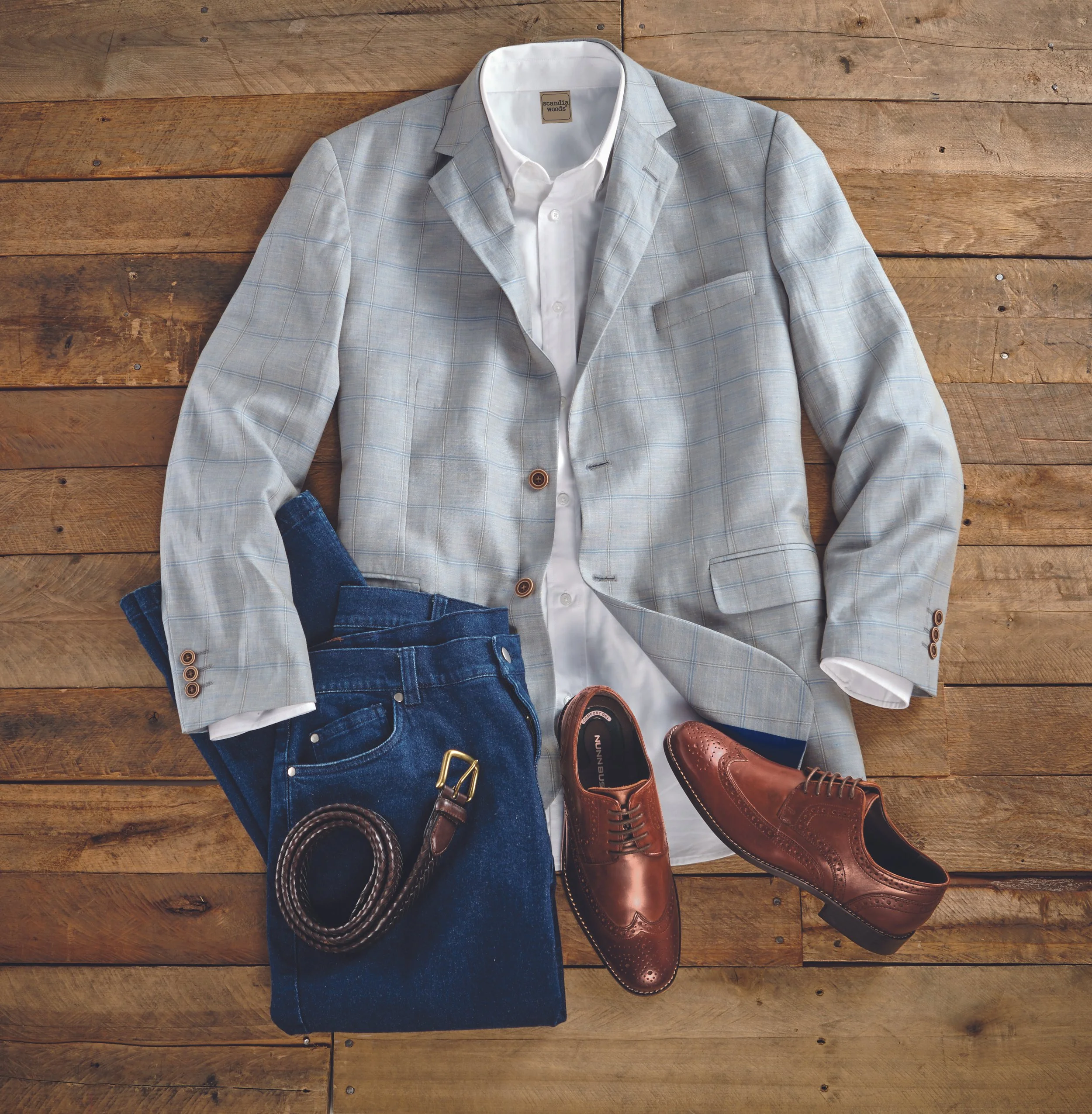 Flat lay of a men's outfit with a light gray plaid blazer, a white dress shirt, dark blue jeans, a black belt, and brown dress shoes on a wooden surface.