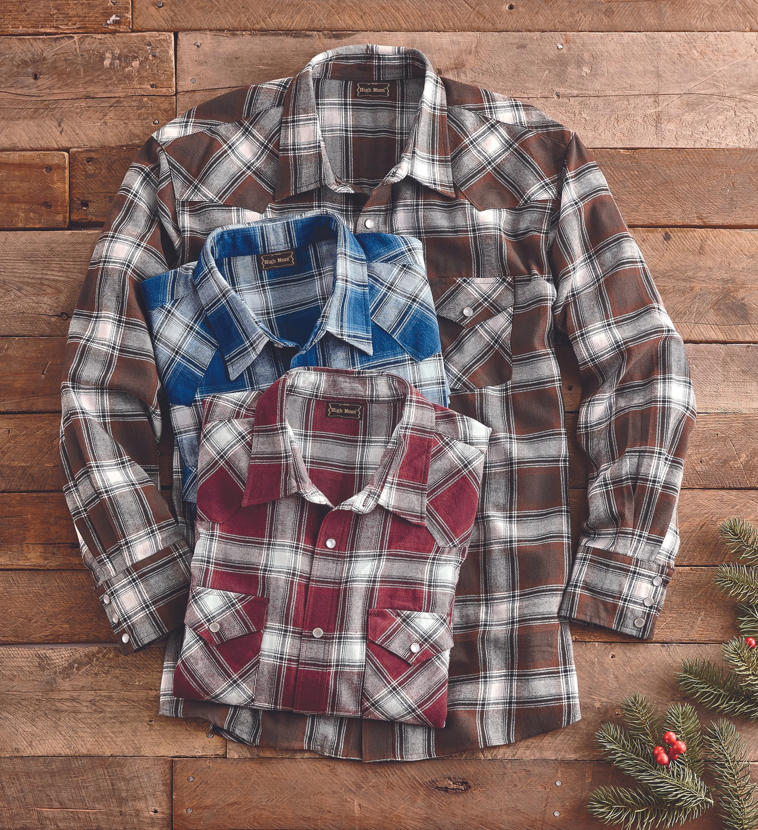 Three plaid flannel shirts in different colors (brown, blue, red) laid on a wooden surface with a Christmas sprig in the lower right corner.