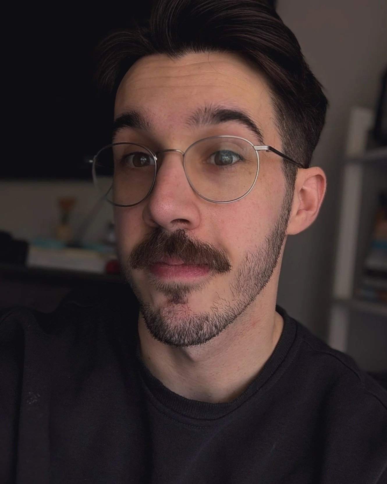 Close-up of a young man with dark hair, glasses, and facial hair, wearing a black shirt, indoors with bookshelves in the background.