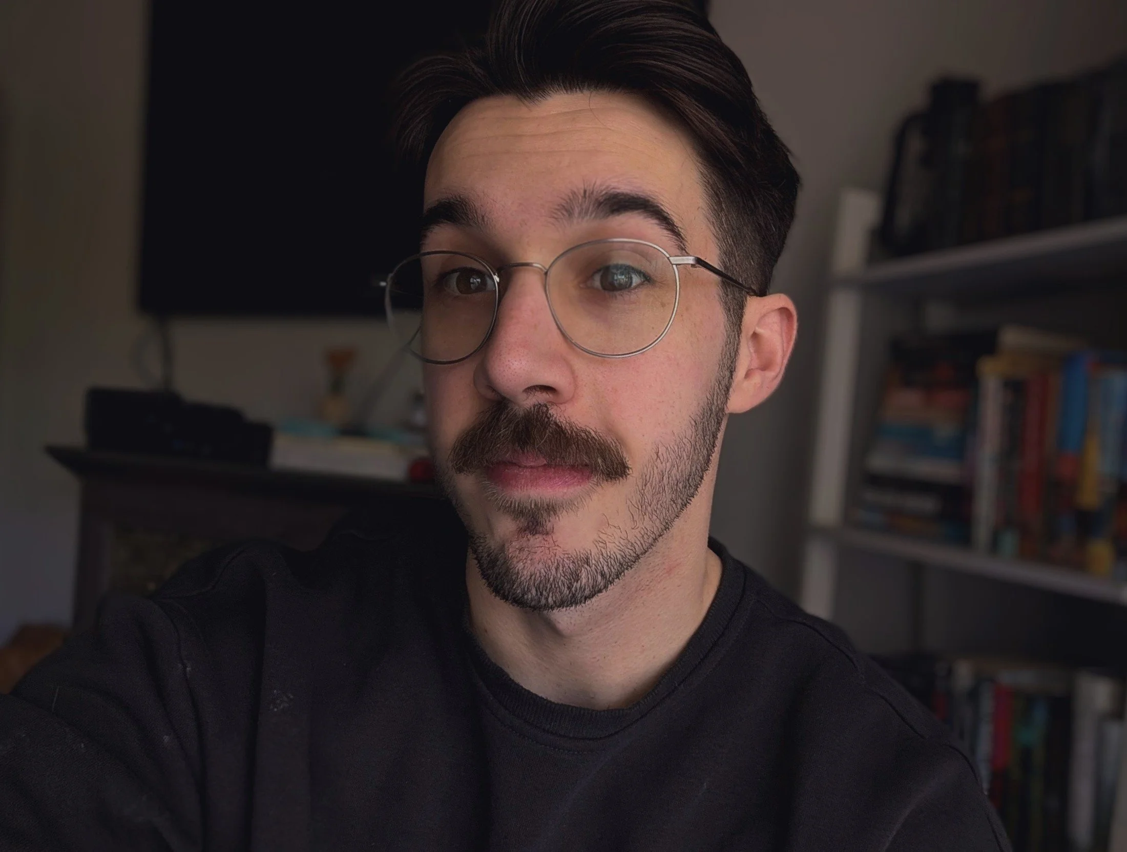 Close-up of a young man with dark hair, glasses, and facial hair, wearing a black shirt, indoors with bookshelves in the background.