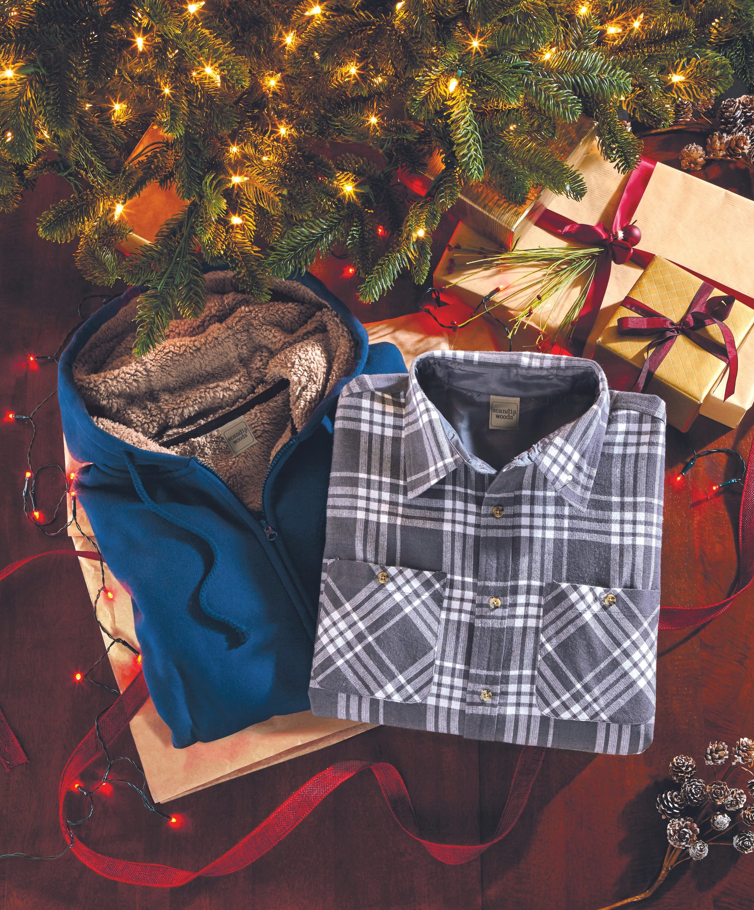 A Christmas scene with a decorated tree, wrapped presents, and shirts laid out on a table, surrounded by pinecones and festive lights.
