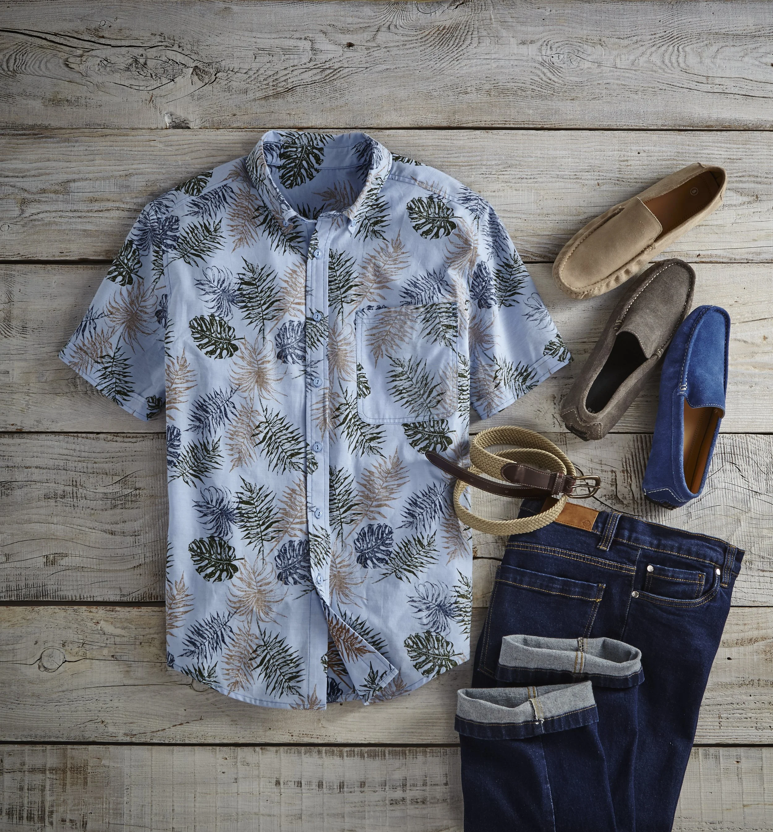 A flat lay of men's fashion items including a short-sleeve button-up shirt with a leafy tropical print, accompanied by a pair of beige loafers, gray loafers, blue loafers, and belts in a neutral and dark blue color, all arranged on a rustic wooden su