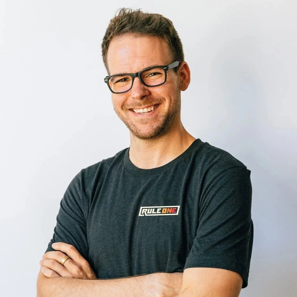 A smiling man with glasses and short brown hair, wearing a black t-shirt with a logo that says 'RULE ONE', standing against a plain white background with arms crossed.