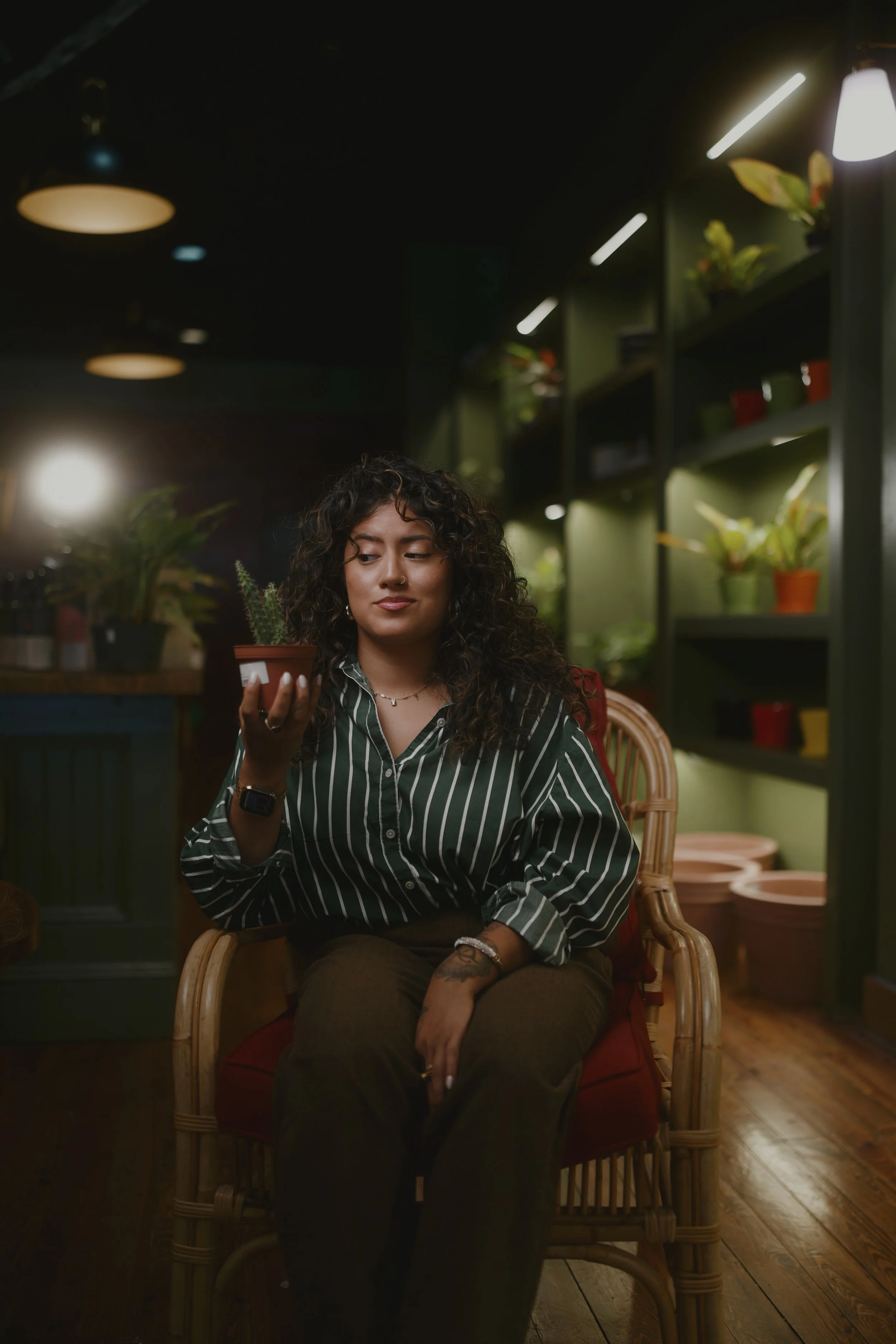 Stephanie Ruiz, founder of A Plant Project, sits in a rattan chair holding a potted plant, surrounded by plant-filled shelves and warm lighting.