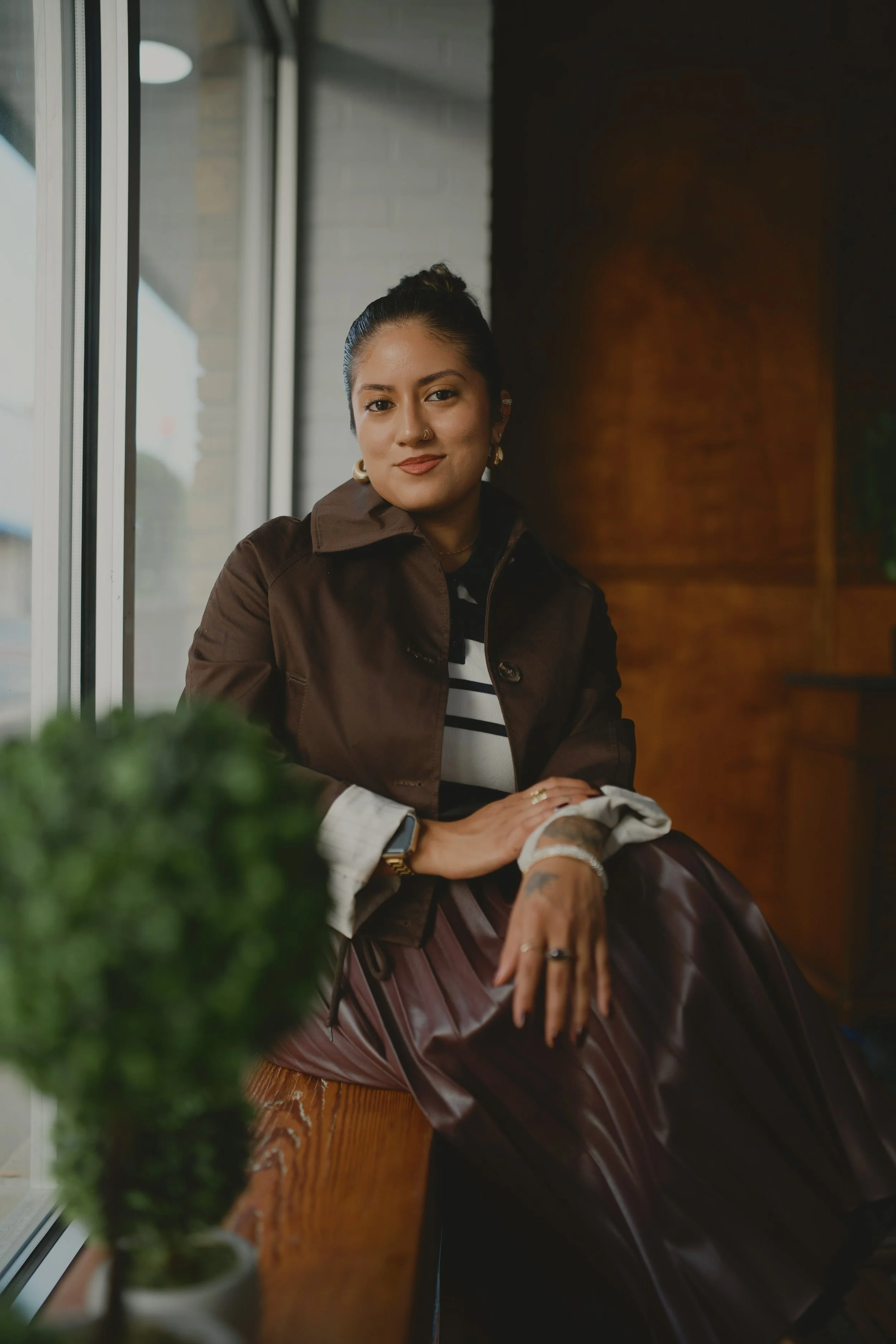 A woman sitting by a window, wearing a brown jacket, striped shirt, and jewelry, with a plant in the foreground and a dark wall behind her.