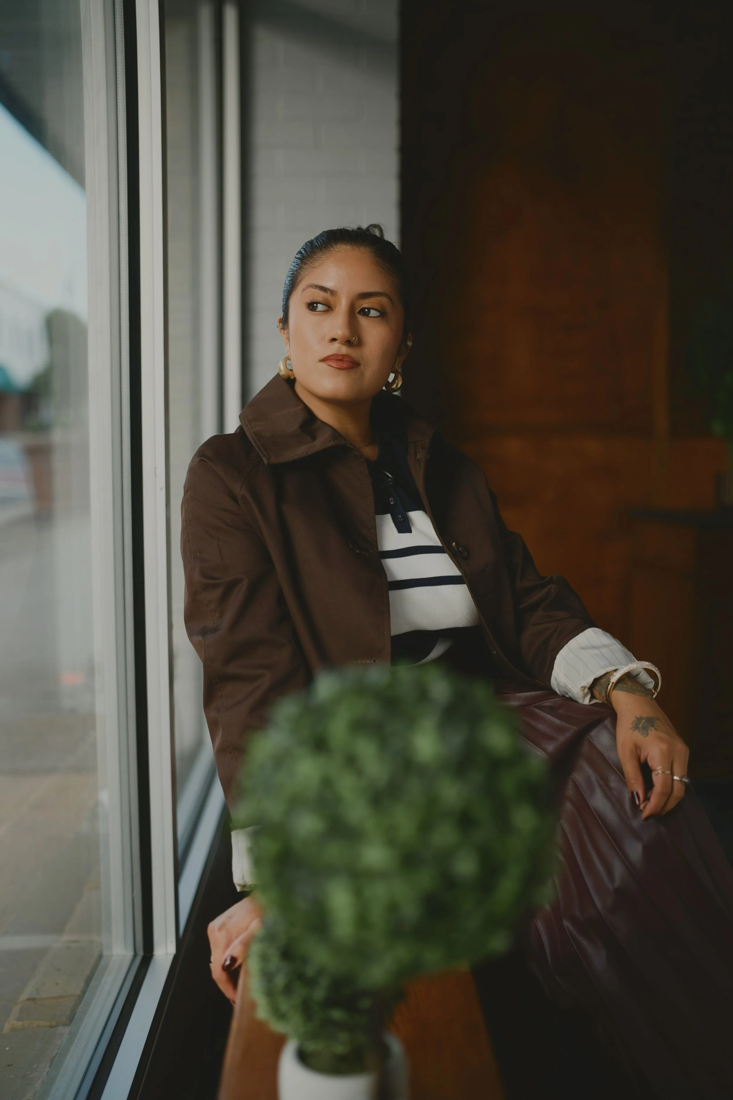 A woman with dark hair tied back, wearing a striped shirt, earrings, and a brown jacket, sitting indoors near a large window with a brick wall behind her, looking to the side with a neutral expression. There is a blurred green plant in the foreground.