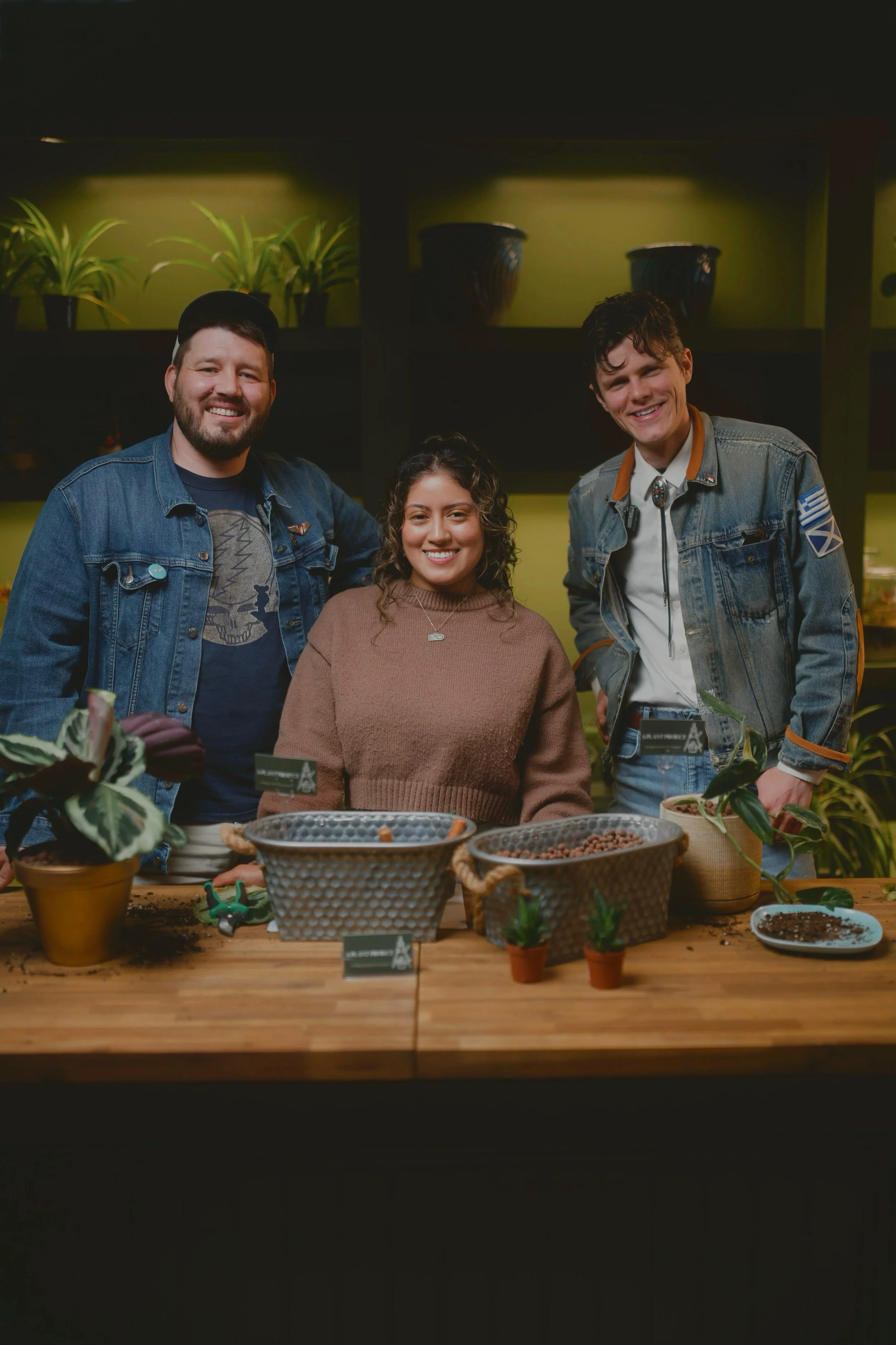 Three people smiling behind a wooden table with plants and small pots, in a well-lit indoor setting.