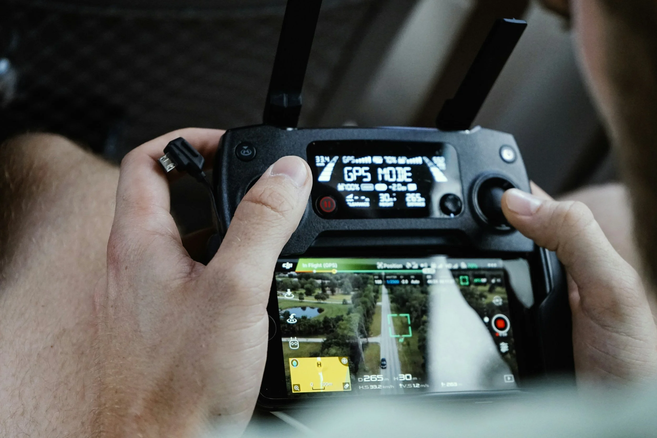 Person holding a drone remote controller with a video feed on a smartphone screen showing a landscape view of trees, roads, and buildings.
