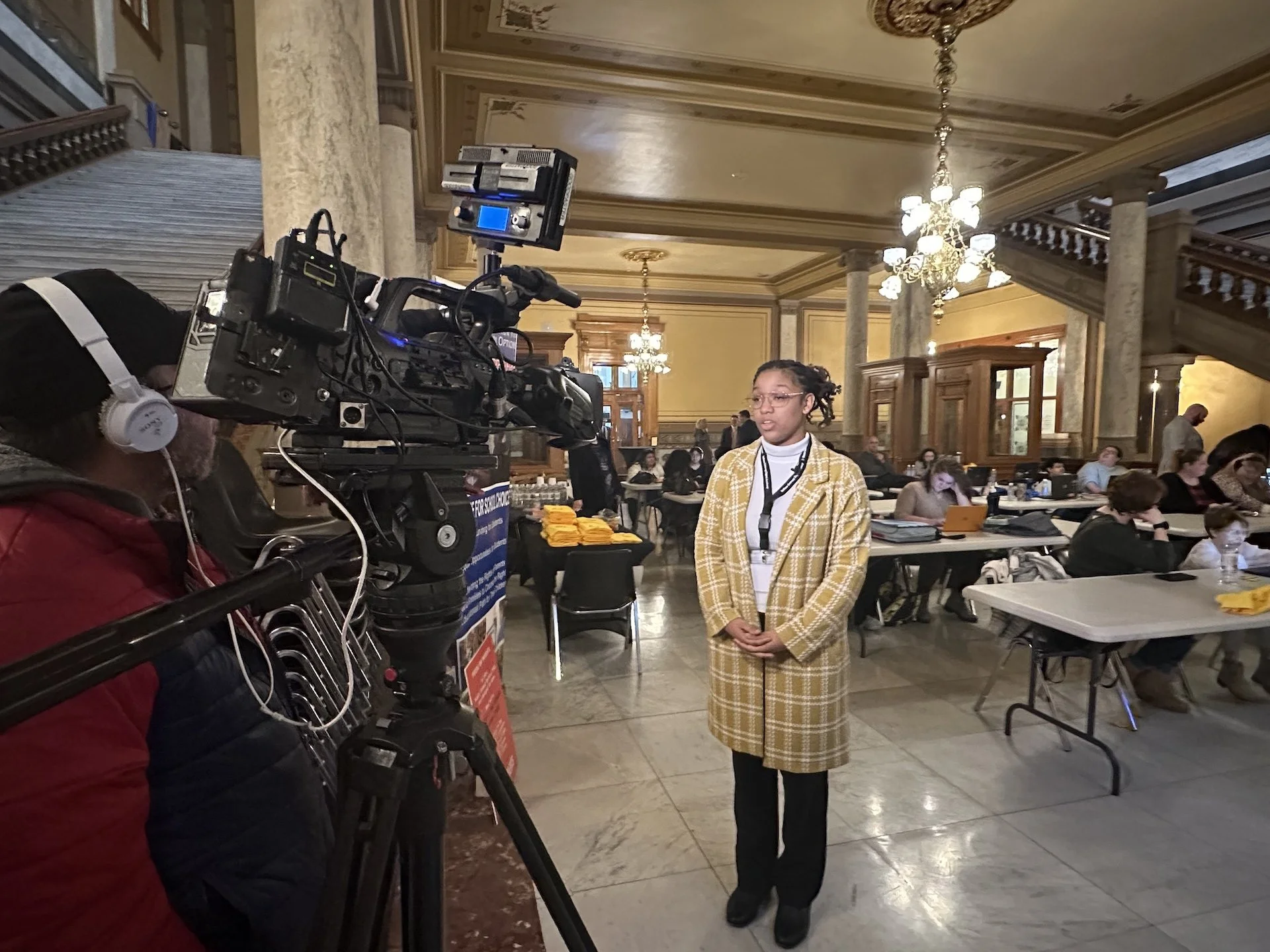 A woman with glasses and a yellow checkered coat standing in front of a camera in a grand, ornate room with chandeliers and many people seated at tables, some using laptops.