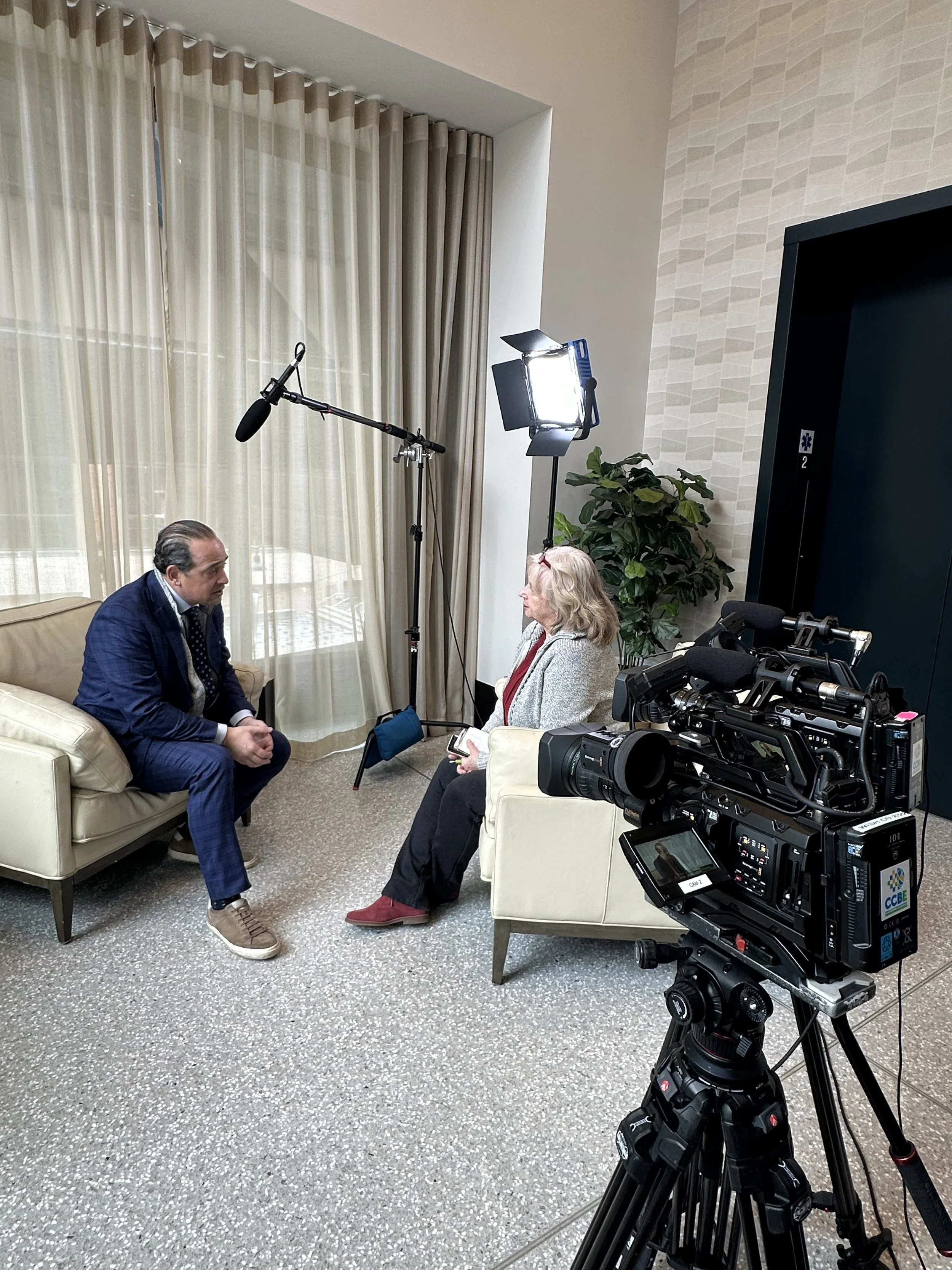 A man and a woman sitting in a hotel room being interviewed or having a conversation with professional video camera equipment and lighting setup.
