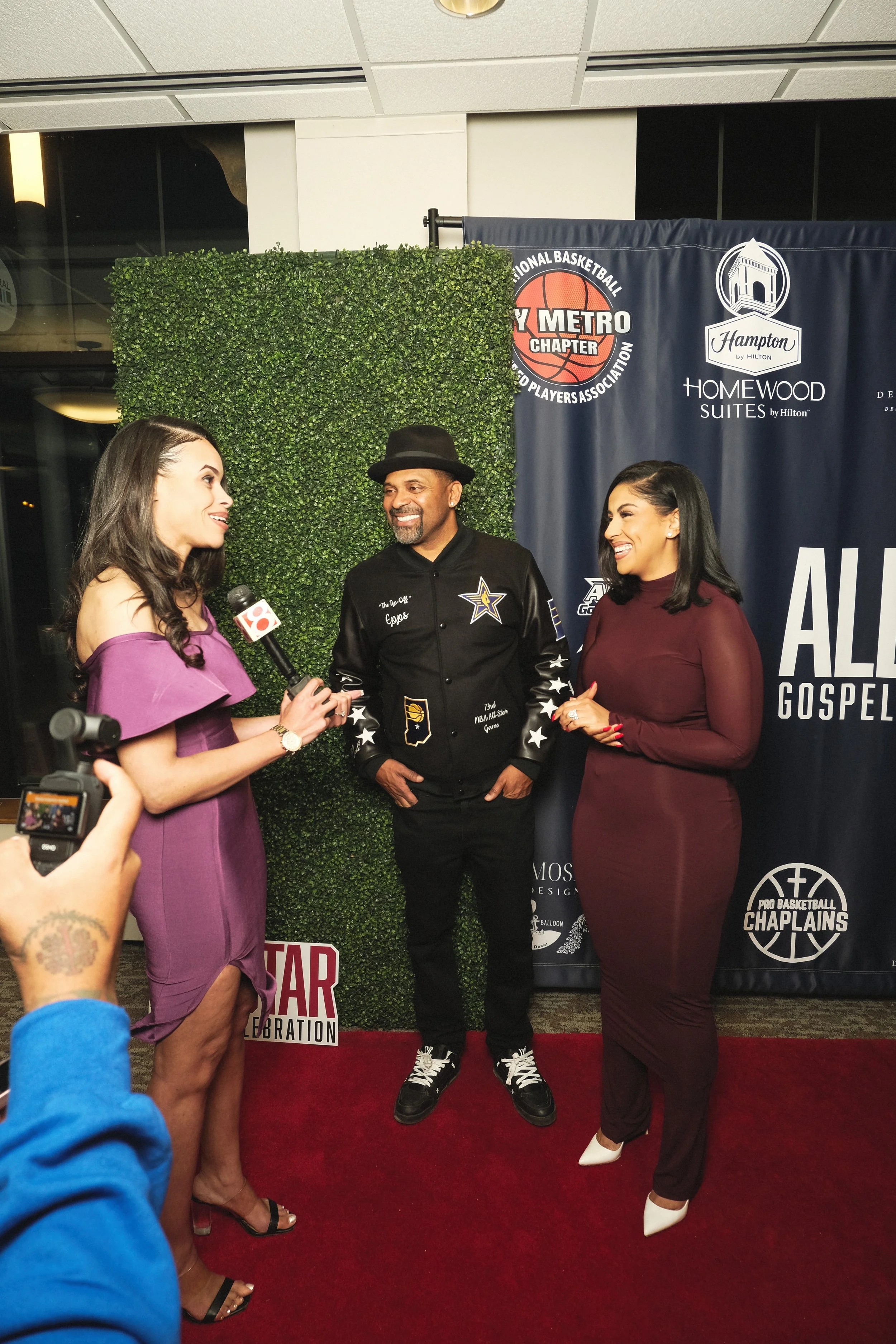 Three women and a man stand on a red carpet at a formal event, smiling and conversing. The woman on the left is holding a microphone, and the woman on the right is wearing a burgundy dress and white shoes. The man in the middle is dressed in a black jacket with a star emblem and a black hat. Behind them is a backdrop with logos and green foliage.