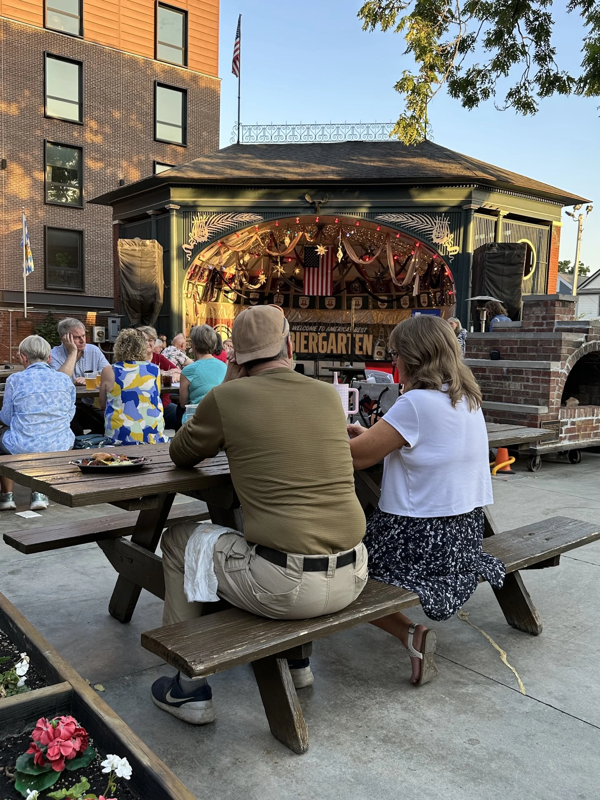 People dining outdoors at a beer garden, with decorated stage and a historical building in the background.