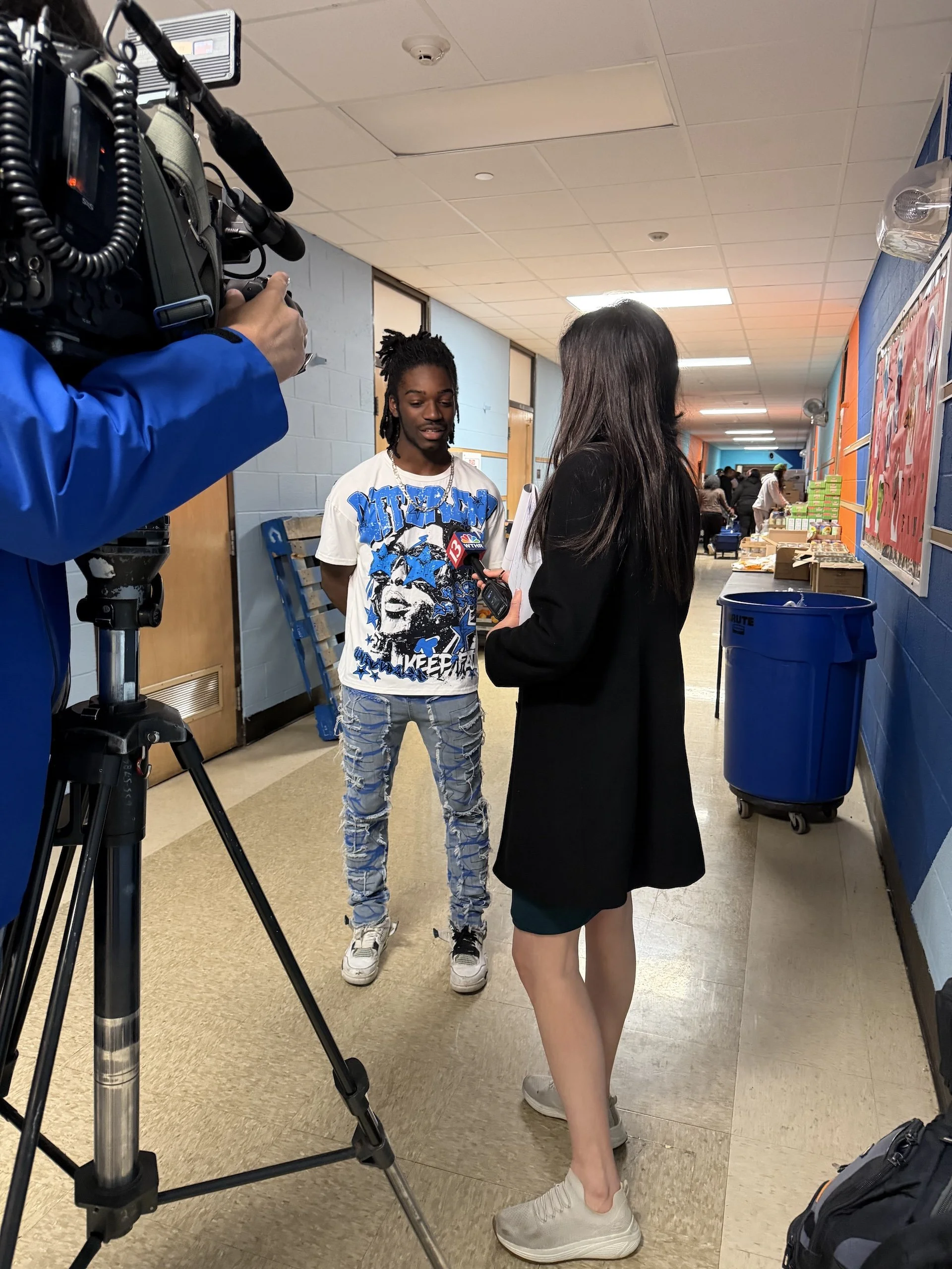 A young man, wearing a graphic t-shirt and distressed jeans, is being interviewed by a woman in a school hallway. A camera and a microphone are pointed at him, and there are people and tables with items in the background.