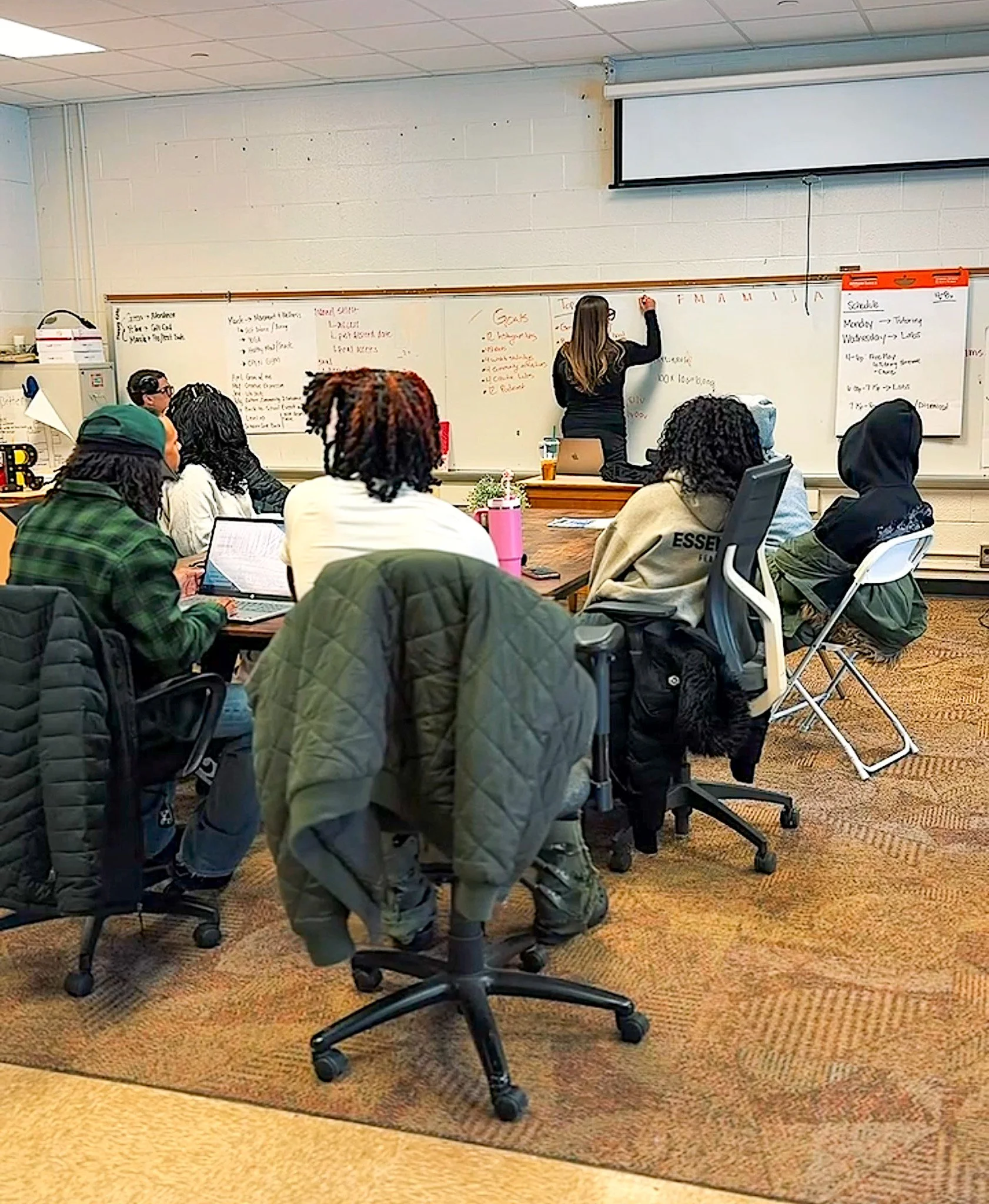 A classroom with students seated around a table, some with laptops and notebooks, listening to a woman writing on a whiteboard. The woman is standing in front of the room, writing on the whiteboard, and classroom supplies are visible on a cabinet to the left.